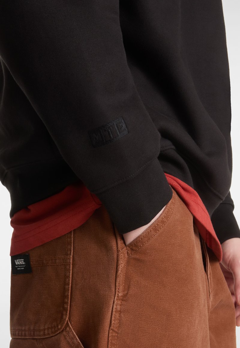 Zwarte sweatshirt met een gestructureerde stof en een reliëflogo op de manchette, gelaagd over een rode shirt en een bruine broek met een label.