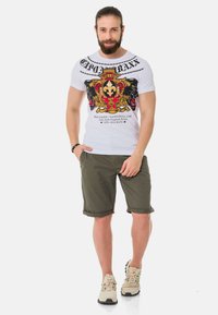 Weißes T-Shirt mit einem bunten Grafikdesign in Gold- und Schwarztönen, kombiniert mit olivgrünen Shorts und beigen Sneakers.