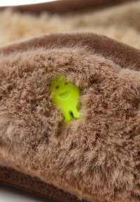 Pantoufle en peluche marron avec une texture douce, présentant une figurine en plastique vert visible émergeant d'un trou dans le matériau.