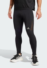 Czarne legginsy sportowe z siateczkowymi wstawkami, dopasowany fason i odblaskowy detal z logo. W zestawie z czarno-białymi butami do biegania.