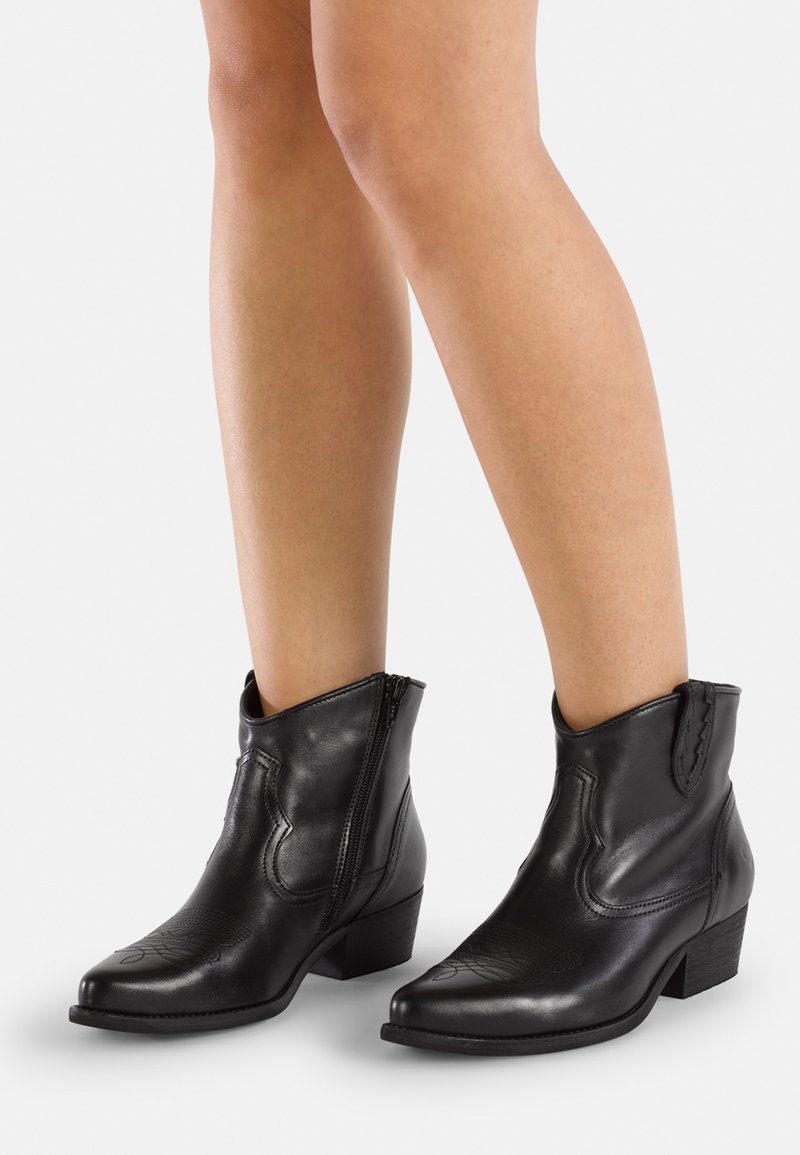 Botas de cuero negras a los tobillos con punta afilada, tacón bajo, cierre de cremallera y costuras decorativas en la parte superior. Textura suave y diseño ajustado.