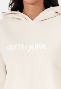 Beżowa bluza z kapturem, wyposażona w kieszeń kangurkę, regulowany kaptur oraz wytłoczony biały napis "SIXTH JUNE PARIS" na klatce piersiowej.