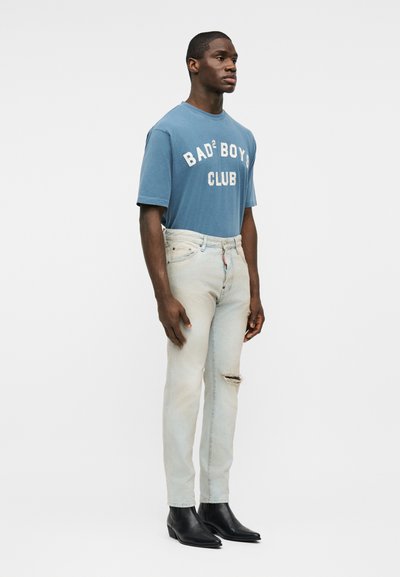 Homme debout portant un t-shirt bleu "Bad Boys Club", un jean clair déchiré et des bottines noires, tourné légèrement vers la droite sur un fond uni.