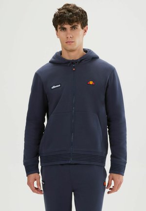 Joven con sudadera con cremallera azul marino de ellesse y pantalones deportivos a juego, posando frente a un fondo claro y liso.