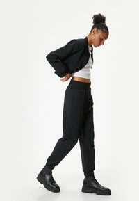 Zwarte bomberjack met drukknoopsluiting, witte cropped top, zwarte joggers met elastische tailleband en chunky zwarte enkellaarzen. Zachte textuur.