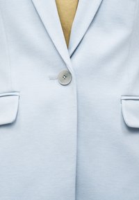 Blazer bleu clair texturé avec un seul bouton gris fermé, comprenant des poches à rabat et une chemise intérieure couleur moutarde visible au niveau du col.