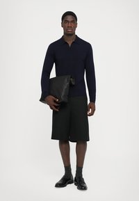 Marinblå stickad polo med krage, svarta skräddarsydda shorts och en slät svart kuvertväska. Svarta läderskor och grå strumpor fulländar looken.