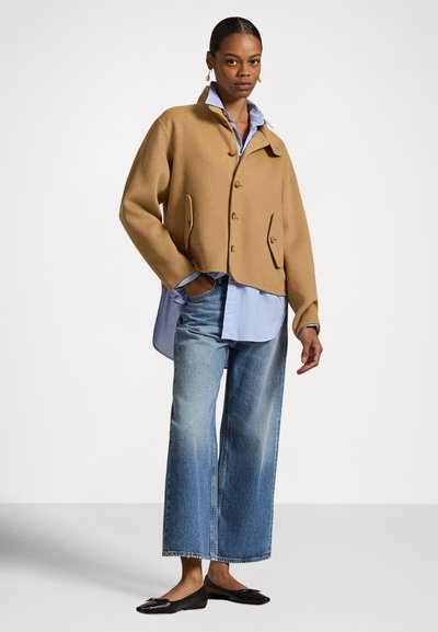 Μικρό μπεζ μάλλινο σακάκι με cropped σχεδιασμό, μεγάλα κουμπιά και πλαϊνές τσέπες, συνδυασμένο με ανοιχτό γαλάζιο πουκάμισο και τζιν παντελόνι φαρδιάς γραμμής.