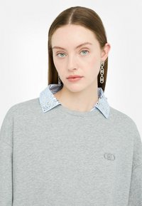 Sweat-shirt gris clair avec un col décoratif rayé bleu présentant un motif clouté, complété par des boucles d'oreilles en chaîne argentée et un logo simple.