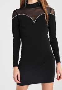 Czarna mini sukienka z długimi rękawami, przezroczystym meshowym dekoltem oraz srebrnymi detalami wzdłuż linii szyi. Gładka tekstura i dopasowany krój.