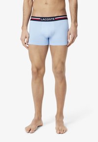 Boxer-briefs bleu clair avec une ceinture rayée noire, rouge et blanche portant la marque "LACOSTE". Tissu en coton doux et extensible.
