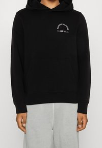 Sudadera de algodón negra con un bolsito frontal, que presenta un logo blanco bordado en el pecho. Puños y dobladillo de canalé, textura suave.
