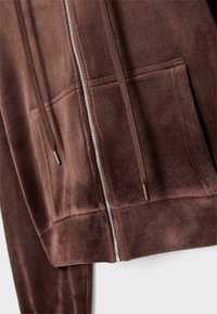 Brązowa bluza z kapturem na zamek, wykonana z miękkiego materiału, z przednim zamkiem błyskawicznym i dwoma kieszeniami z przodu z sznurkami. Gładka tekstura, casualowy design.