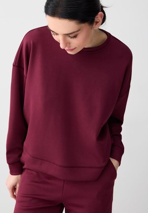 Femme portant un sweat-shirt bordeaux ample à manches longues et un pantalon assorti, regardant vers le bas avec une main dans la poche.