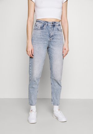 Person wearing light blue high-waisted jeans, white cropped top, white socks, and white sneakers, standing against plain light background.