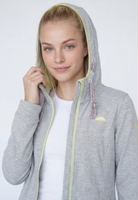 Mladá žena s blond vlasmi má na sebe sivú mikinu na zips s ružovými a bielymi šnúrkami, cez bielu košeľu, jemne sa usmieva.