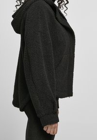 Veste en polaire noire texturée avec capuche, manches larges et coupe oversize, présentant une surface douce et un design court.