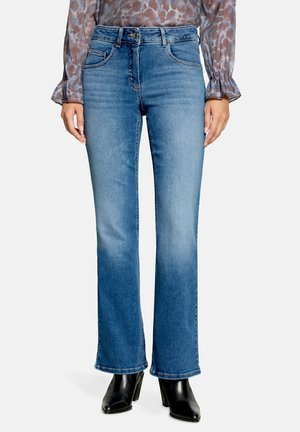 Kvinde iført blå bootcut jeans, en langærmet mønstret bluse og sorte ankelstøvler med hæl, stående mod en ensfarvet hvid baggrund.