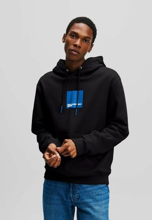 Jonge man die een zwarte hoodie met blauw vierkant logo en blauwe spijkerbroek draagt, staand tegen een effen lichte achtergrond, kijkend naar de camera.