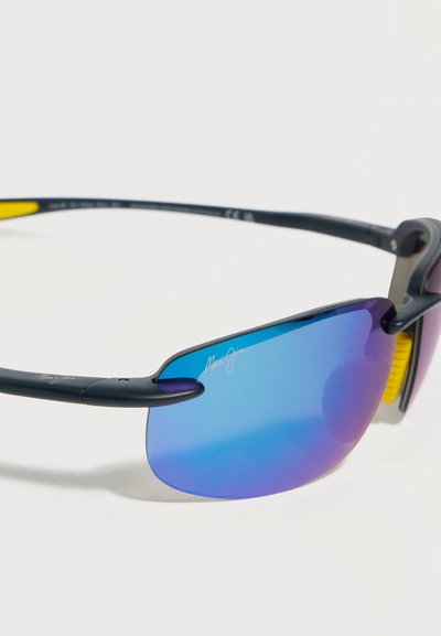 Αθλητικά γυαλιά ηλίου με μπλε φακούς με καθρέφτη, μαύρο πλαίσιο και κίτρινα μαξιλαράκια στη μύτη, σε γωνία που δείχνει τη σήμανση στο πλάι και την καμπυλότητα των φακών.