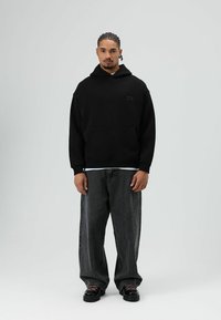 Schwarzer Hoodie mit einer Fronttasche und kleinem Logo, kombiniert mit locker sitzenden grauen Denim-Jeans. Schwarze Schuhe mit pinkfarbenen Schnürsenkeln.