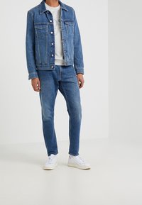 Mann in blauer Jeansjacke, blauen Jeans, grauem Sweatshirt und weißen Sneakern, der vor einem schlichten weißen Hintergrund steht.