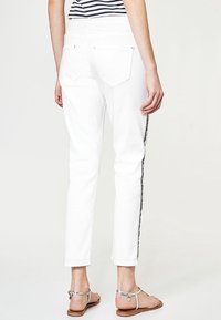 Bílé slim fit džíny s černým bočním pruhem, dvěma zadními kapsami a nerovným lemem. Modelka má na sobě páskové sandály.