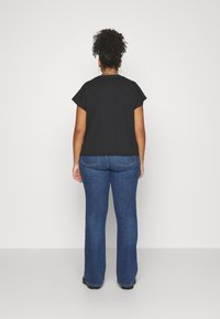 Femme aux cheveux bouclés portant un t-shirt noir et un jean bleu, se tenant face à un mur gris clair uni, les mains détendues sur les côtés.