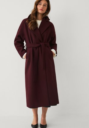 Woman wearing a long burgundy belted coat with large lapels, hands in pockets, white sweater sleeves visible, and black heels.