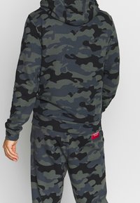 Haut à capuche camouflage dans des tons de vert foncé, gris et noir avec une texture douce. Comprend un écusson logo au dos et une capuche pour une protection supplémentaire.