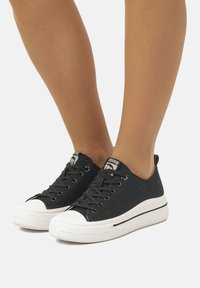 Zapatillas de lona negras con suelas de goma blanca, punteras redondas y cordones negros. Los patrones en la suela proporcionan agarre. Logo visible en la lengüeta.