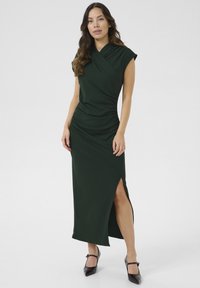 Robe enveloppe verte foncée à manches courtes, avec fronces sur les côtés et une haute fente, fabriquée dans un tissu lisse et extensible. Chaussures noires à bout pointu.