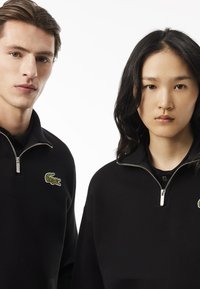 Sudaderas negras con cremallera que presentan una textura acanalada, con un logo de cocodrilo verde en una y un logo azul en la otra. Cuellos con cremallera y ajuste casual.