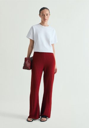 Mujer con camiseta blanca de manga corta, pantalones rojos texturizados de talle alto, sandalias negras, sosteniendo un bolso tejido de color rojo oscuro, de pie contra un fondo liso.