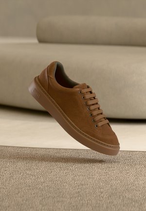 Brauner Wildleder-Sneaker mit passenden Schnürsenkeln schwebt über einem Teppichboden, beiges Sofa im Hintergrund.