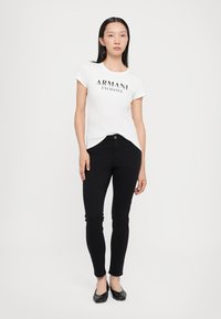 Balta īsām piedurknēm T-kreklu ar "ARMANI EXCHANGE" logotipu, apvienojot ar melnajām pieguļošajām džinsām un melnām dzīvokļu kurpēm uz neitrāla fona.