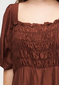 Rudos spalvos raukšlėtas audinys su pūstomis trumpomis rankovėmis ir šiek tiek raukšlėtu kvadratiniu iškirpimu ant žmogaus.