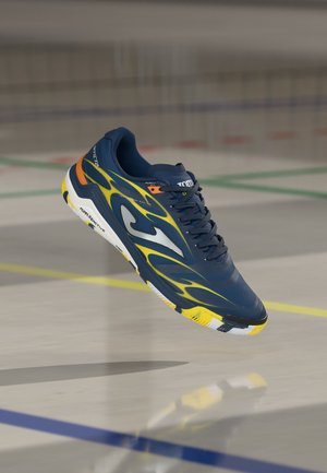 Marineblauer Sportschuh mit gelb-orange Flammendesign, weiße und gelbe Sohle, geschnürte Schnürsenkel, schwebt über dem Boden eines Indoor-Sportplatzes.