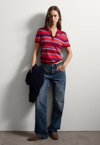 Rotes Poloshirt mit weißen und blauen Streifen, kombiniert mit blauen Jeans. Das Modell hält einen marineblauen Pullover und trägt braune Slipper.