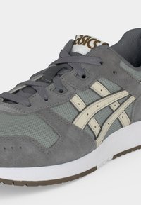 Γκρι αθλητικό παπούτσι Asics με κρεμ ριγέ, πλέγμα και σουέντ επάνω μέρος, γκρι κορδόνια και λευκή σόλα με καφέ πέλμα.