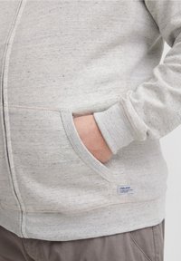 Hånden delvis inne i frontlommen på en lys grå zip-up hettegenser med en liten etikett på kanten av lommen.
