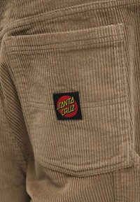 Μπεζ υφασμάτινη κορντούρα με κάθετες ρίγες, που διαθέτει μια μαύρη ετικέτα με το κόκκινο και κίτρινο λογότυπο "Santa Cruz" στη δεξιά τσέπη.