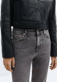 Chaqueta corta de cuero negra con un acabado texturizado, combinada con jeans grises de talle alto que tienen remaches de cobre y bolsillos clásicos.