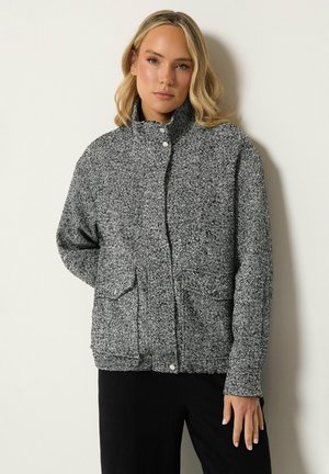Femme blonde portant une veste à motif chevrons noir et blanc avec boutons-pression et poches, debout contre un mur clair uni.