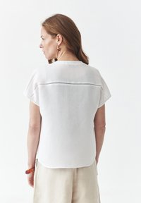 Hvid linnedtop med korte ærmer, rund halsudskæring og diskret detaljer på ryggen. Bæres med beige linnedbukser.