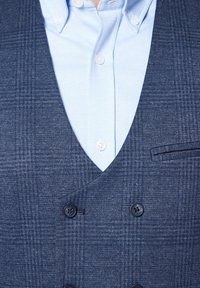 Granatowy, dwurzędowy kamizelka z teksturowanym wzorem w pieszą kratę, z dwoma czarnymi guzikami i kieszenią, noszona na jasnoniebieskiej koszuli w warkocz.