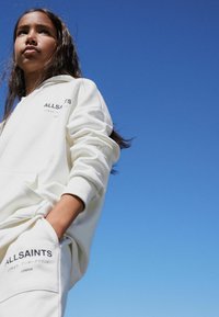 Krem dukserica s kapuljačom i crnim logotipom "ALLSAINTS". Jednostavan dizajn, mekani materijal, dugi rukavi. U pozadini se vidi vedro plavo nebo.