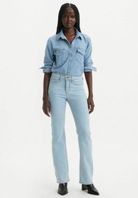 Jeans de mezclilla de color azul claro combinados con una camisa de botones a juego, con bolsillos en el pecho y mangas enrolladas, complementados con botas negras hasta el tobillo.