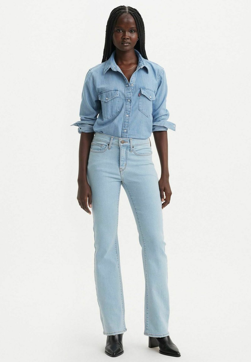 Jeans de mezclilla de color azul claro combinados con una camisa de botones a juego, con bolsillos en el pecho y mangas enrolladas, complementados con botas negras hasta el tobillo.