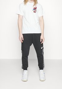 Valkoinen puuvillainen t-paita, jossa on värikäs graafinen kuvio rinnassa, yhdistettynä mustiin collegehousuihin, joissa on sivutaskut ja valkoiset Nike-logo-akkentssit.